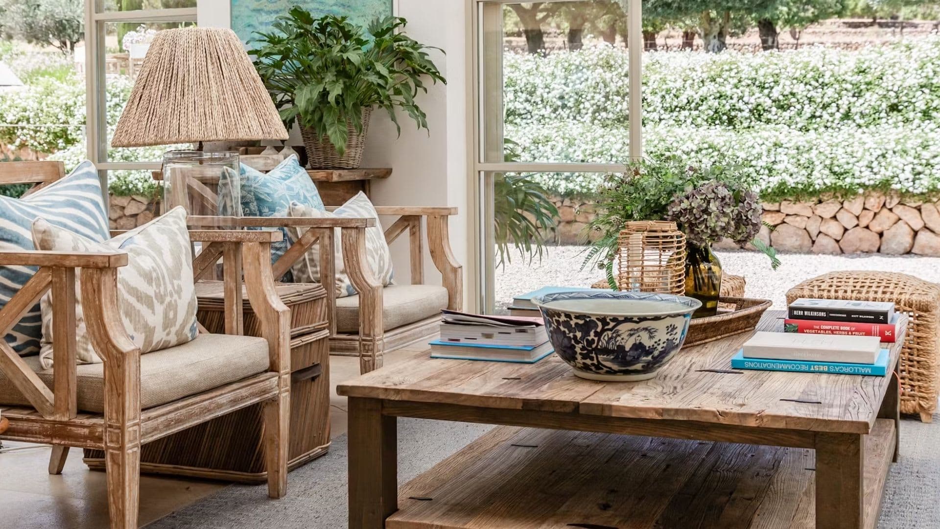 A sitting room in rattan and bleached wood, full-height windows open onto a flowering garden