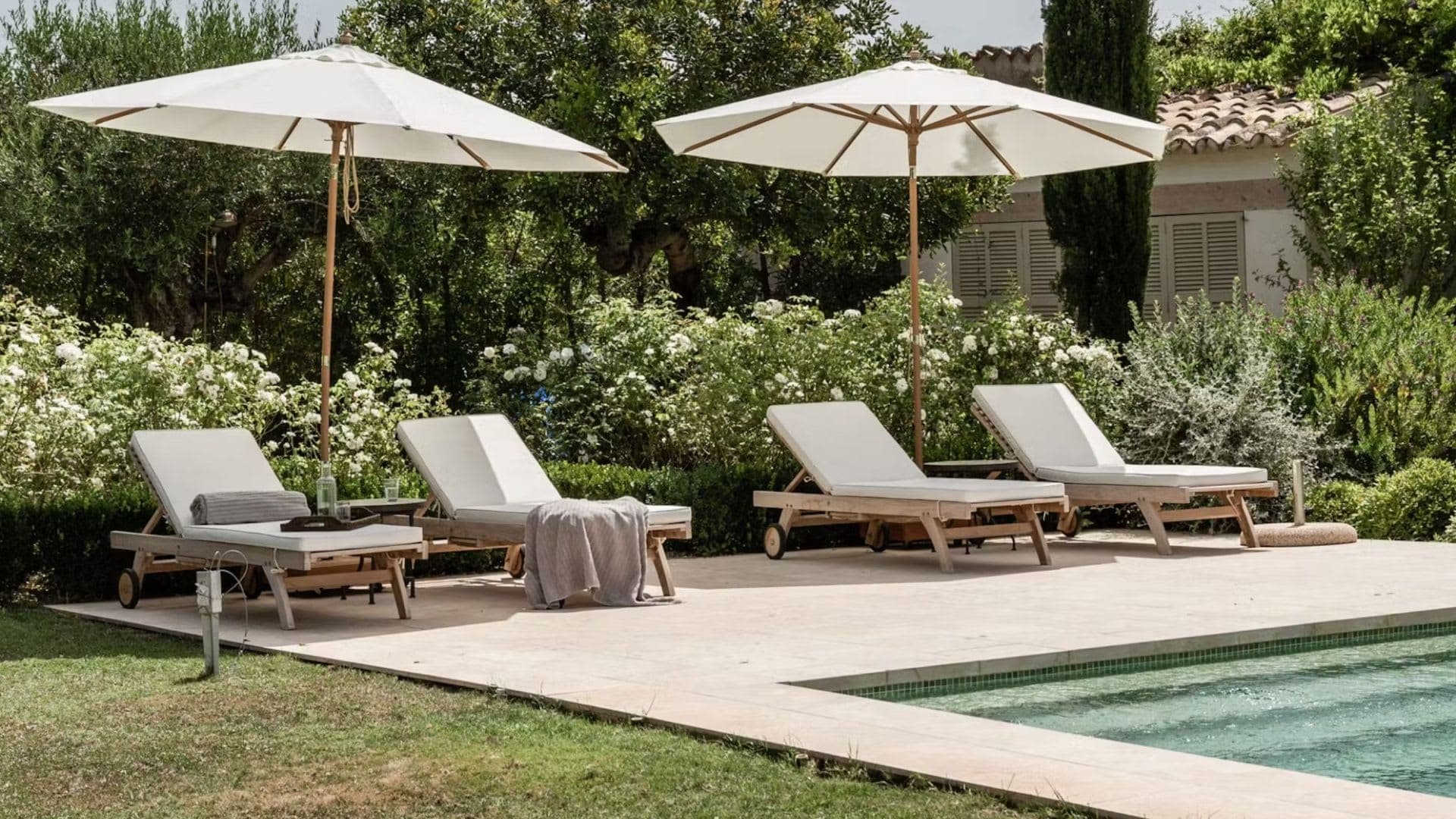 Four sun loungers under cream parasols on a stone terrace beside a pool, white roses behind