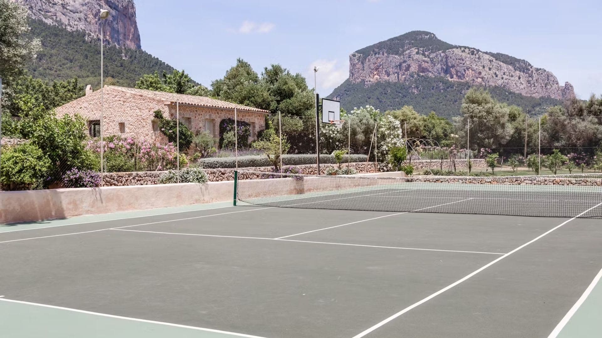 A tennis court beside a stone farmhouse, with mountains behind